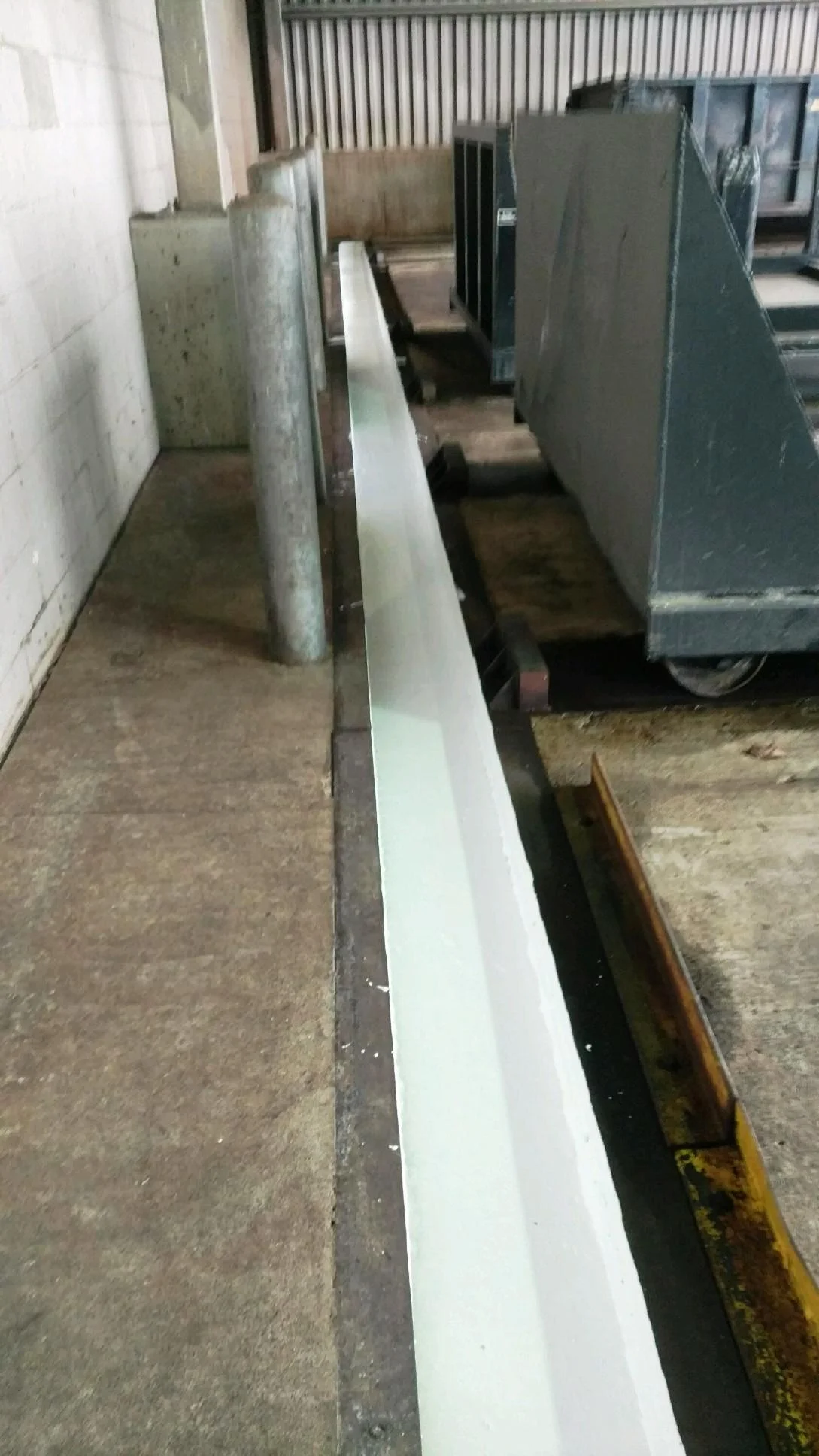 A long, rectangular, light-colored gutter in an industrial setting with a steel column and machinery.