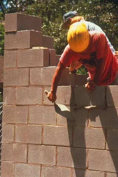 Construction worker laying cement blocks, applying mortar with a trowel, wearing a hard hat.