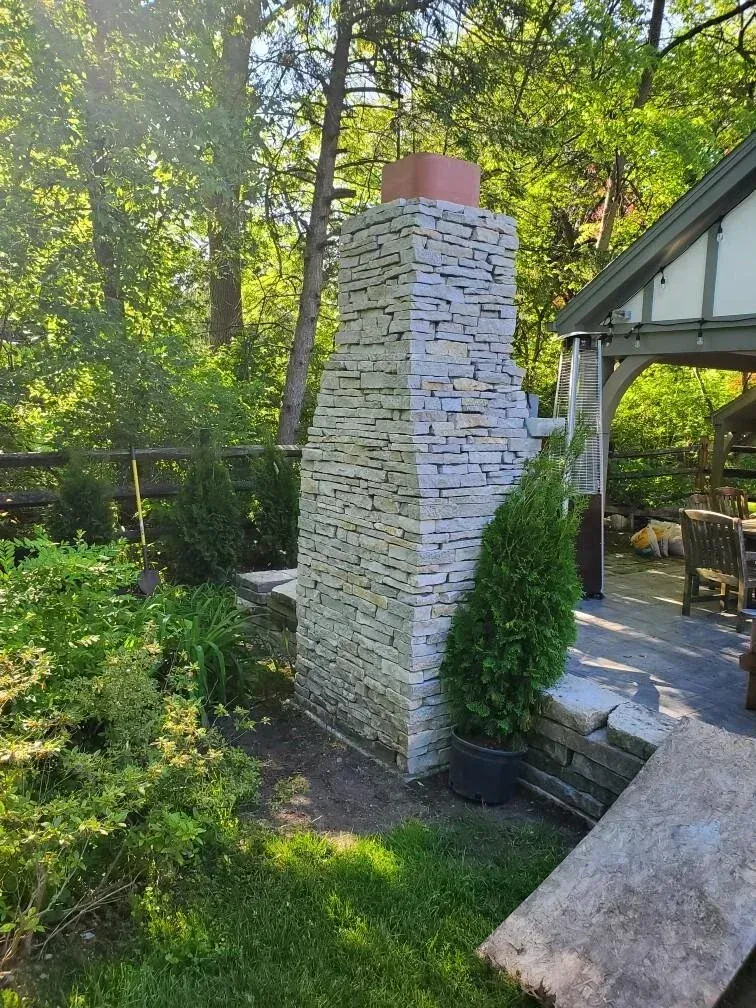 Stone chimney outdoors with copper top, beside a patio. Lush green foliage and trees in background.