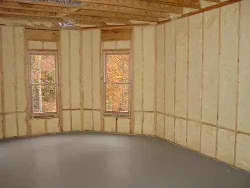Interior of a room with spray foam insulation between wooden studs, two windows, and a gray floor.