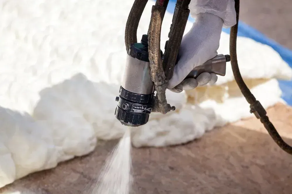 A worker in gloves sprays expanding foam insulation onto a surface.