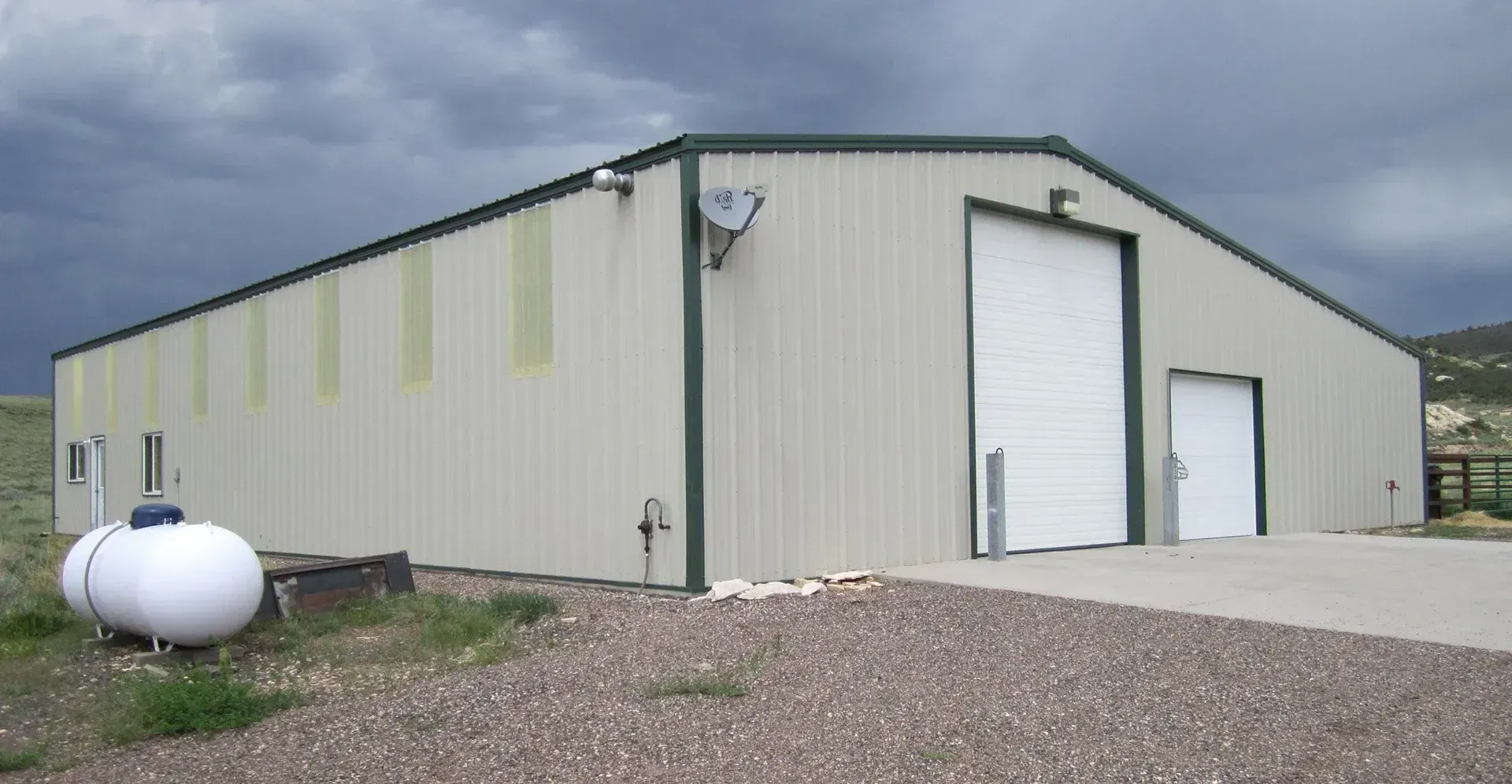 Tan metal building with green trim, large garage door, propane tank, under a cloudy sky.