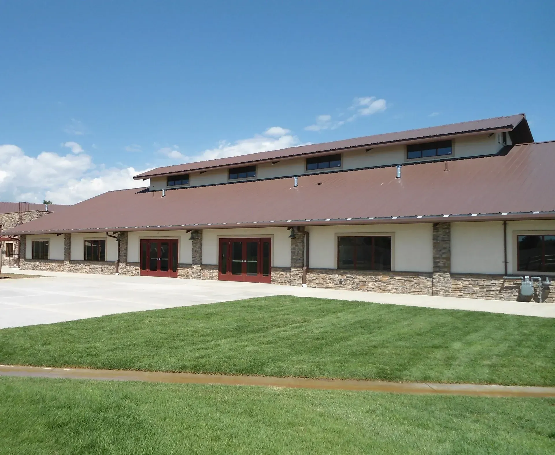 Brown-roofed building with stone accents, windows, and red doors. Green lawn in the foreground under a blue sky.