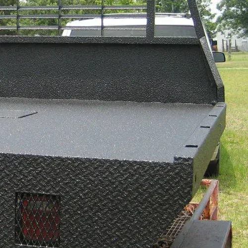 Black, textured truck bed with metal rack, tail light visible. Green grass and car in background.