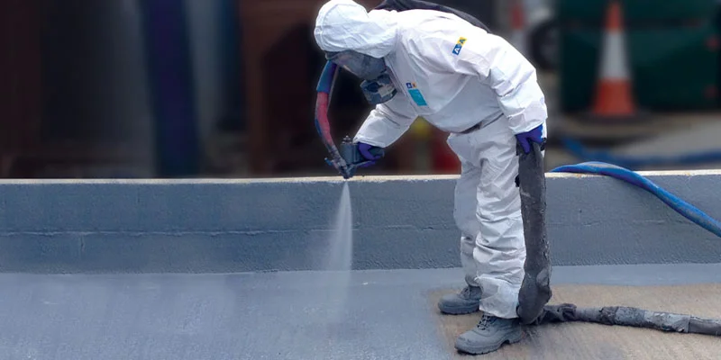 Person in protective suit spraying a surface with a substance, likely paint or sealant. Outdoor setting.