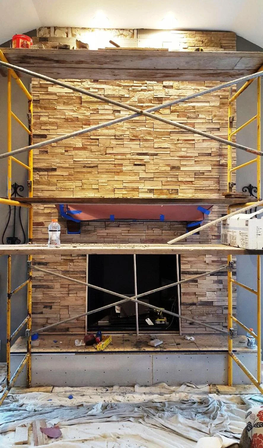 Fireplace under construction, stone veneer, scaffolding, tools, and a partial wooden mantel.