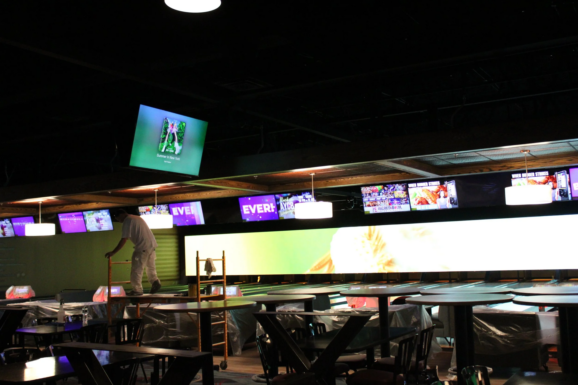 Bowling alley interior with multiple TVs displaying sports, a person on a ladder, tables, and chairs.