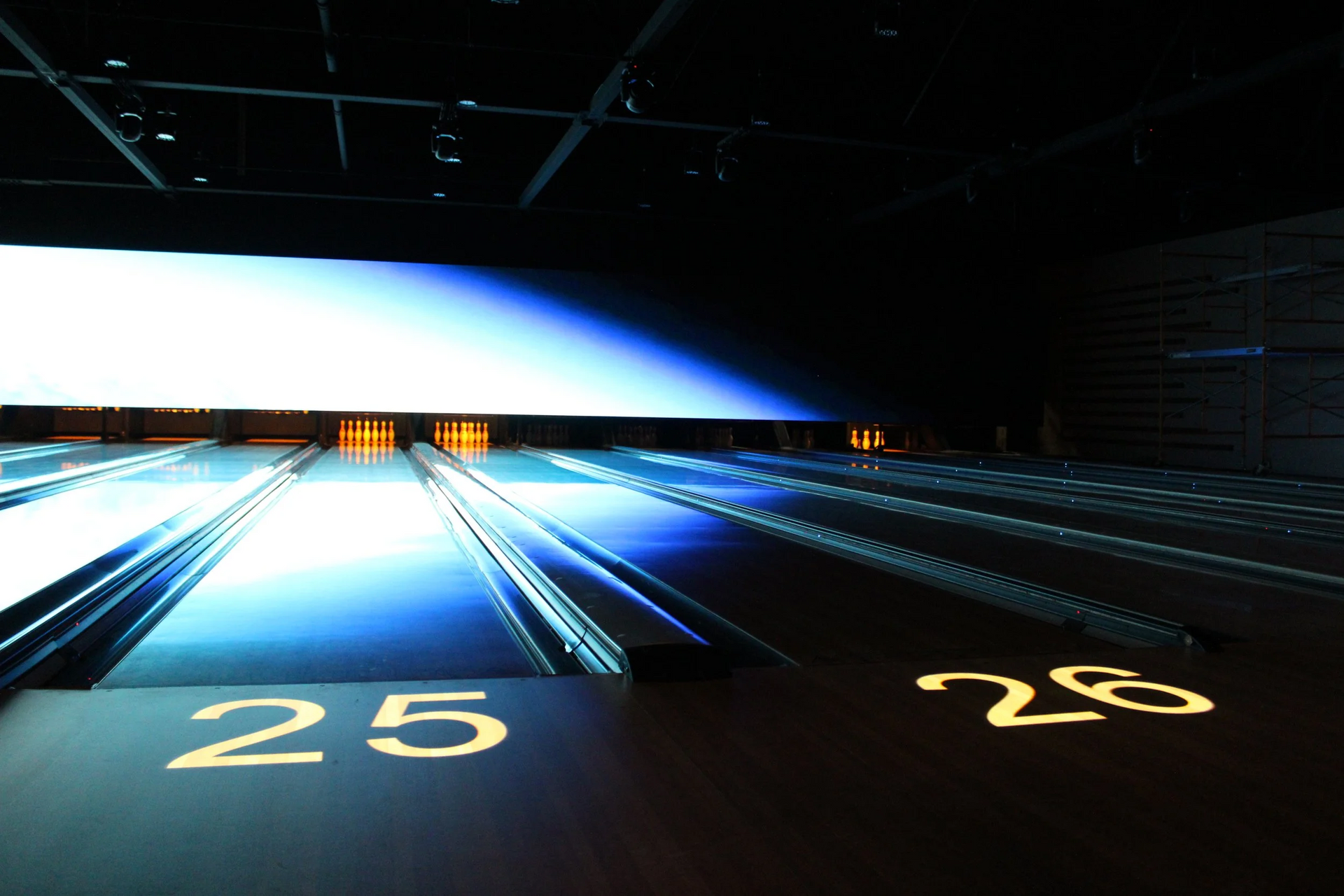 Bowling lanes 25 and 26 lit with blue and white lights, dark interior, illuminated overhead screen.