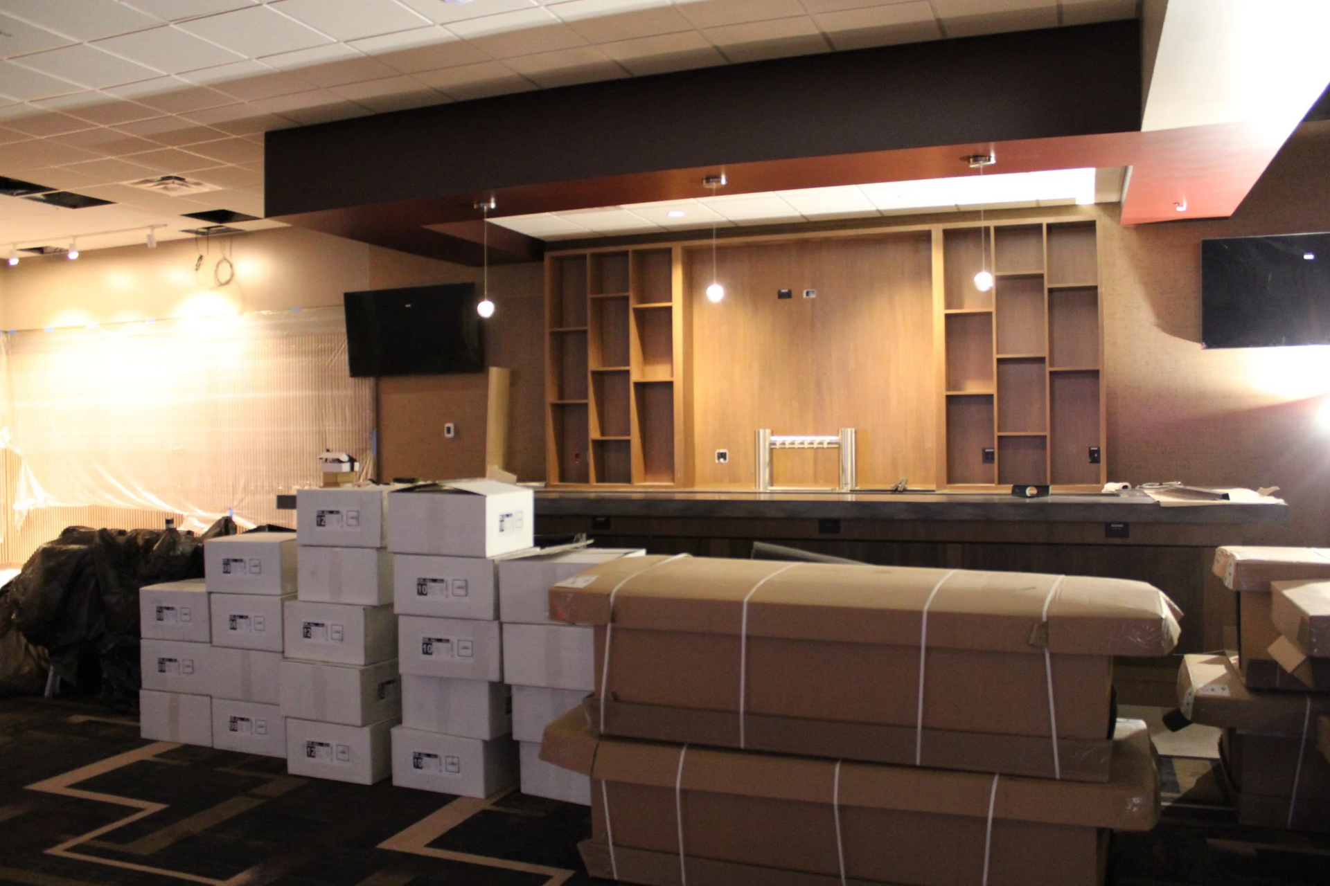 Boxes and wrapped furniture in a room, with a bar, shelves, and TVs; interior undergoing construction.
