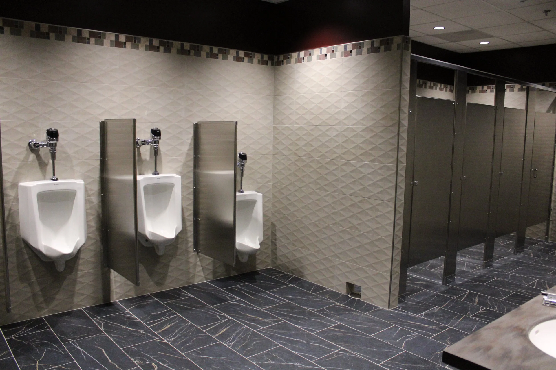 Public restroom with urinals and stalls. Gray and white tiled walls, dark flooring.