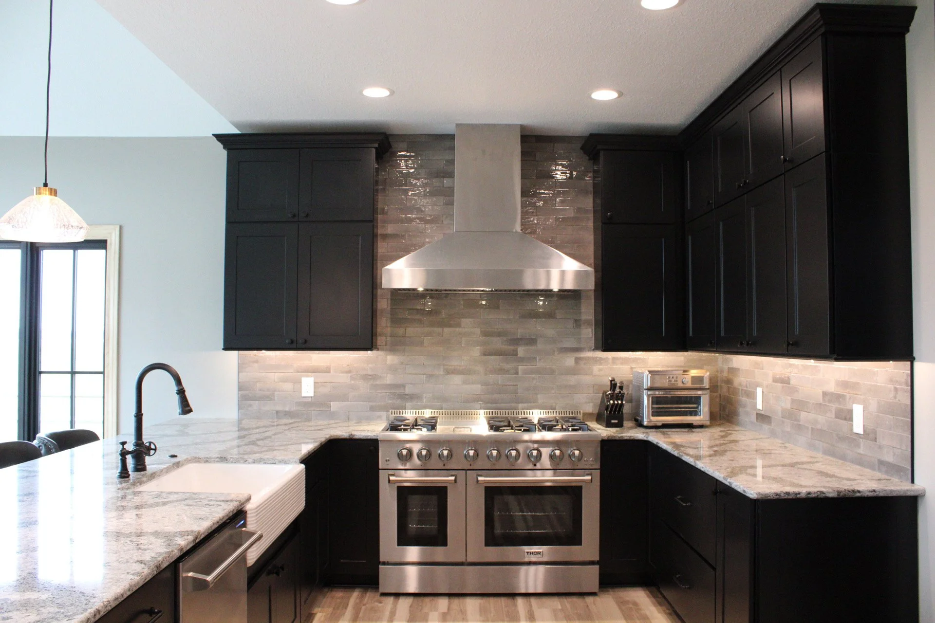 Black cabinets in a modern kitchen with stainless steel appliances, a gray backsplash, and a light-colored countertop.