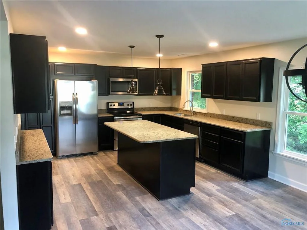Modern kitchen with black cabinets, stainless steel appliances, granite counters, and gray wood-look flooring.
