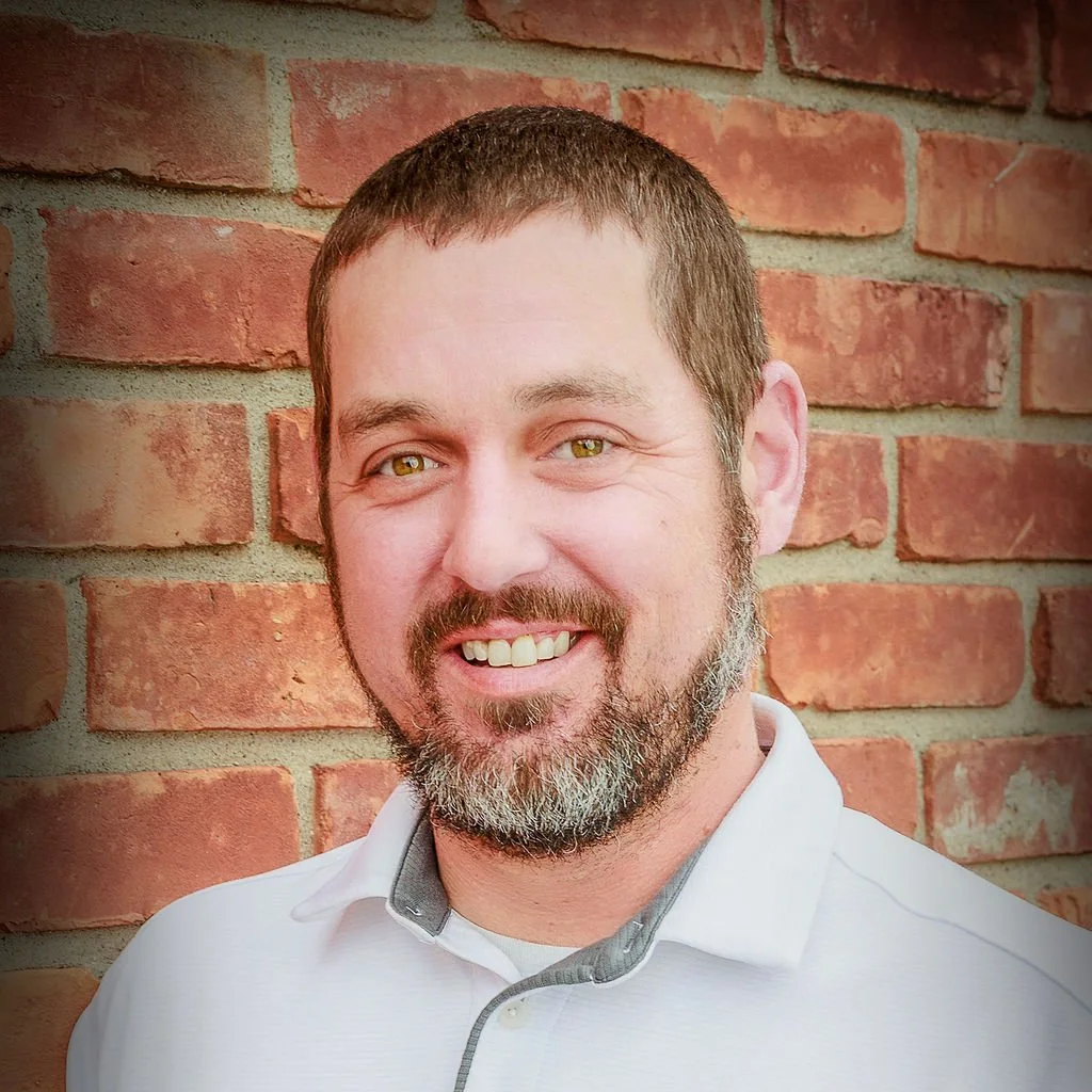 Man with a beard smiles in front of a brick wall, wearing a white collared shirt.