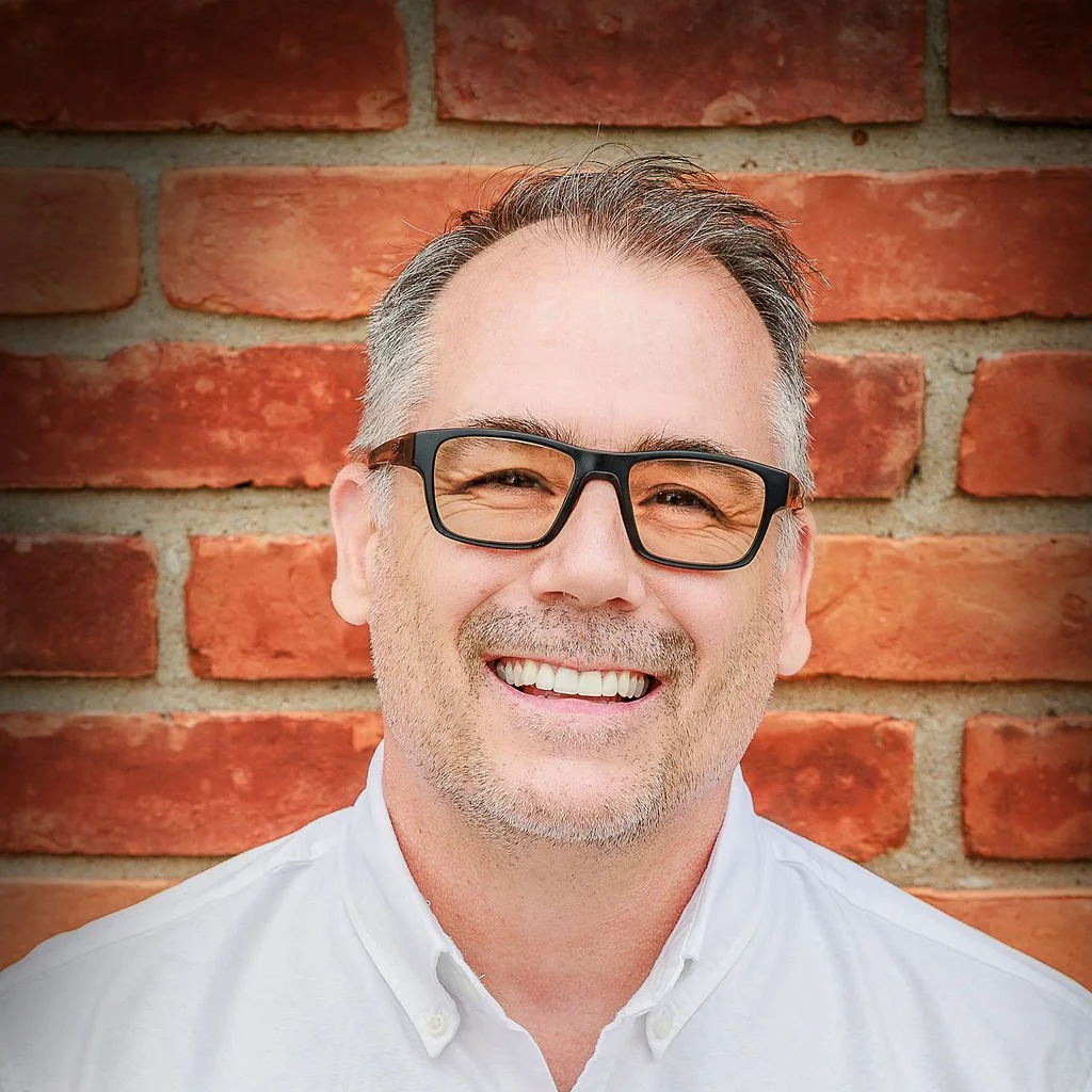 Man with glasses smiles in front of a brick wall. He is wearing a white shirt.