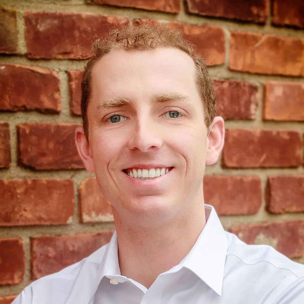 Smiling man with fair skin and red hair, wearing a white shirt, in front of a brick wall.