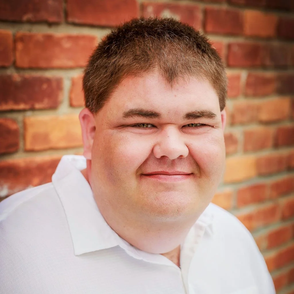 Smiling man with short brown hair in white shirt against brick wall.