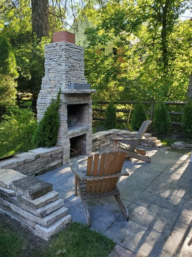 Outdoor fireplace with seating on a stone patio, surrounded by trees and greenery.