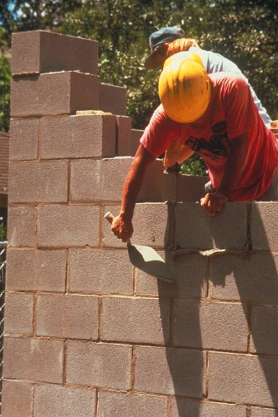 A construction worker in a yellow helmet lays concrete blocks, applying mortar with a trowel.