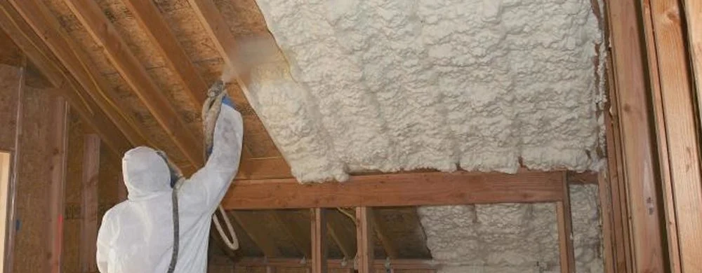 A person in protective gear sprays foam insulation onto a wooden ceiling in a home's attic.