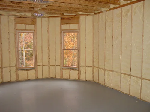 Interior view of a room with spray foam insulation between wooden studs. Windows offer a view of trees.