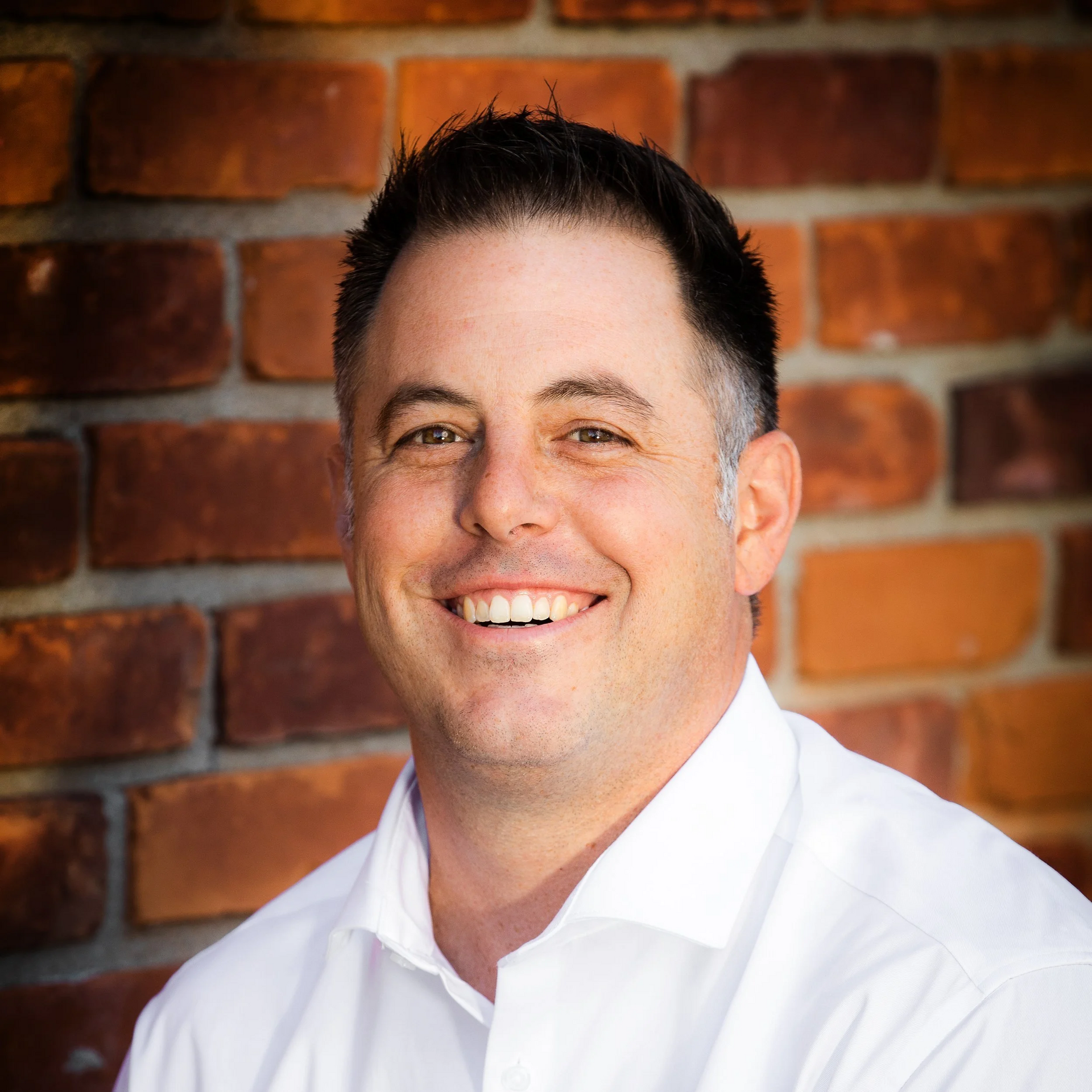 Man with short dark hair smiles, wears a white shirt, in front of a brick wall.