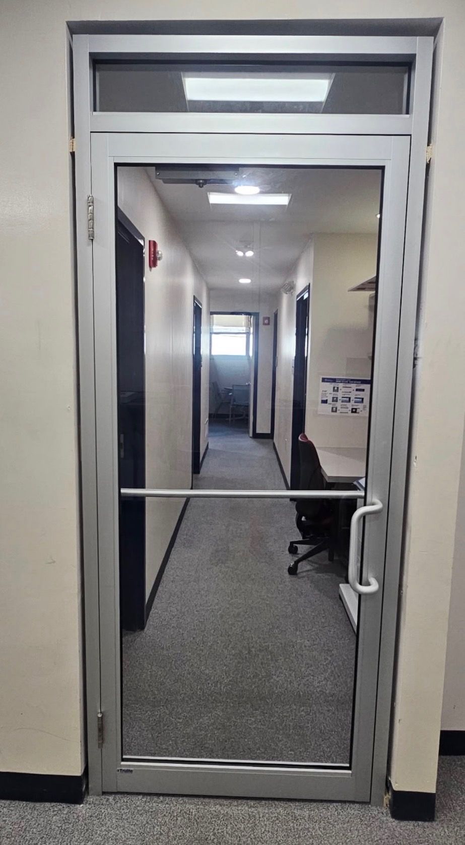 Glass office doorway opening to a carpeted hallway with ceiling lights and a chair inside.