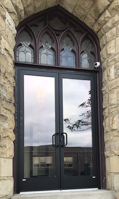 Dark double doors with arched windows above, set in a stone building. Reflecting sky and nearby building.