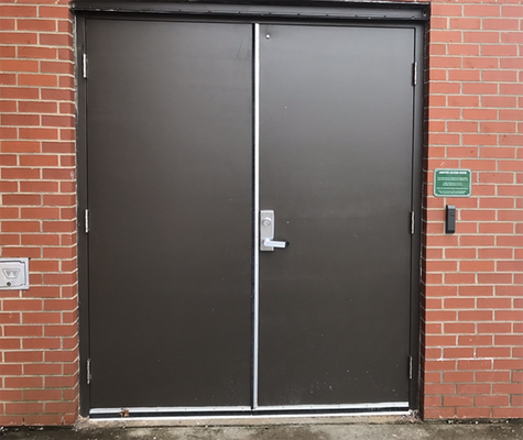 Double dark gray metal doors set in a brick wall with a silver handle and a small sign to the right.