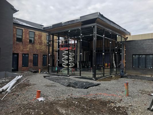 Construction site: Glass-walled structure attached to brick building. Exterior work in progress, cloudy sky.