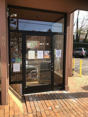 Black door entrance to a business with three signs, brick floor, and window above the door.