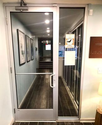 Glass door opens into a long hallway with framed art, wood flooring, and recessed lighting. A sign hangs on the inside.