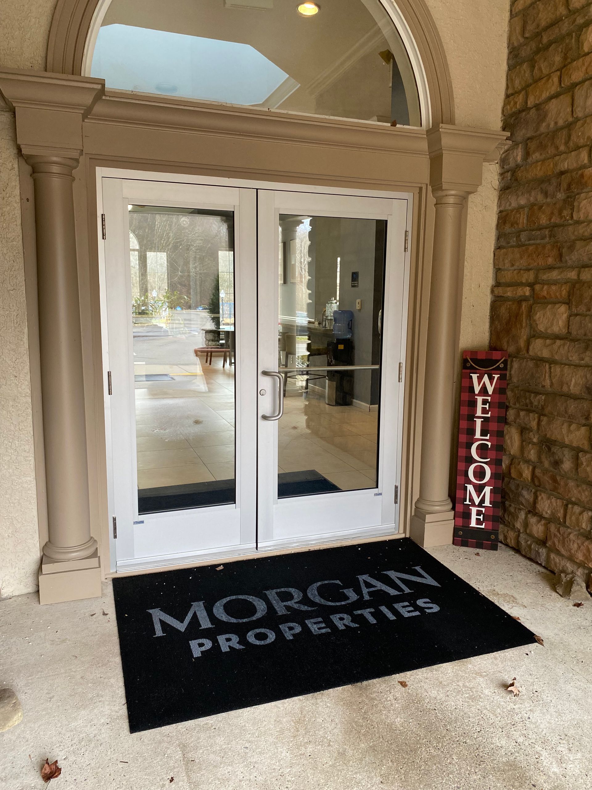 Double glass doors at a Morgan Properties building entrance with a branded doormat and a red buffalo check welcome sign.