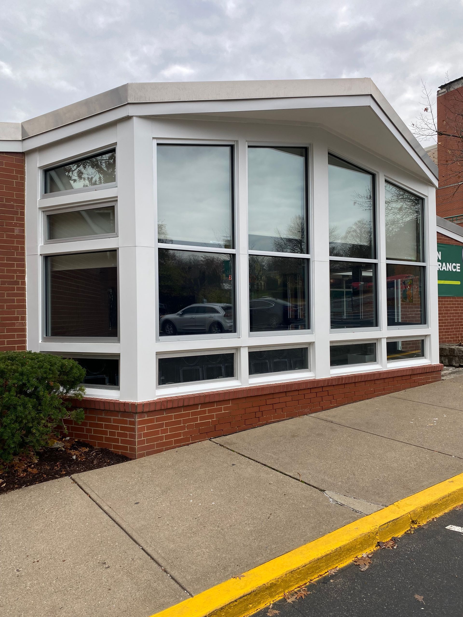 Exterior view of a building with a white-framed bay window, a red brick base, and a paved sidewalk with a yellow curb.