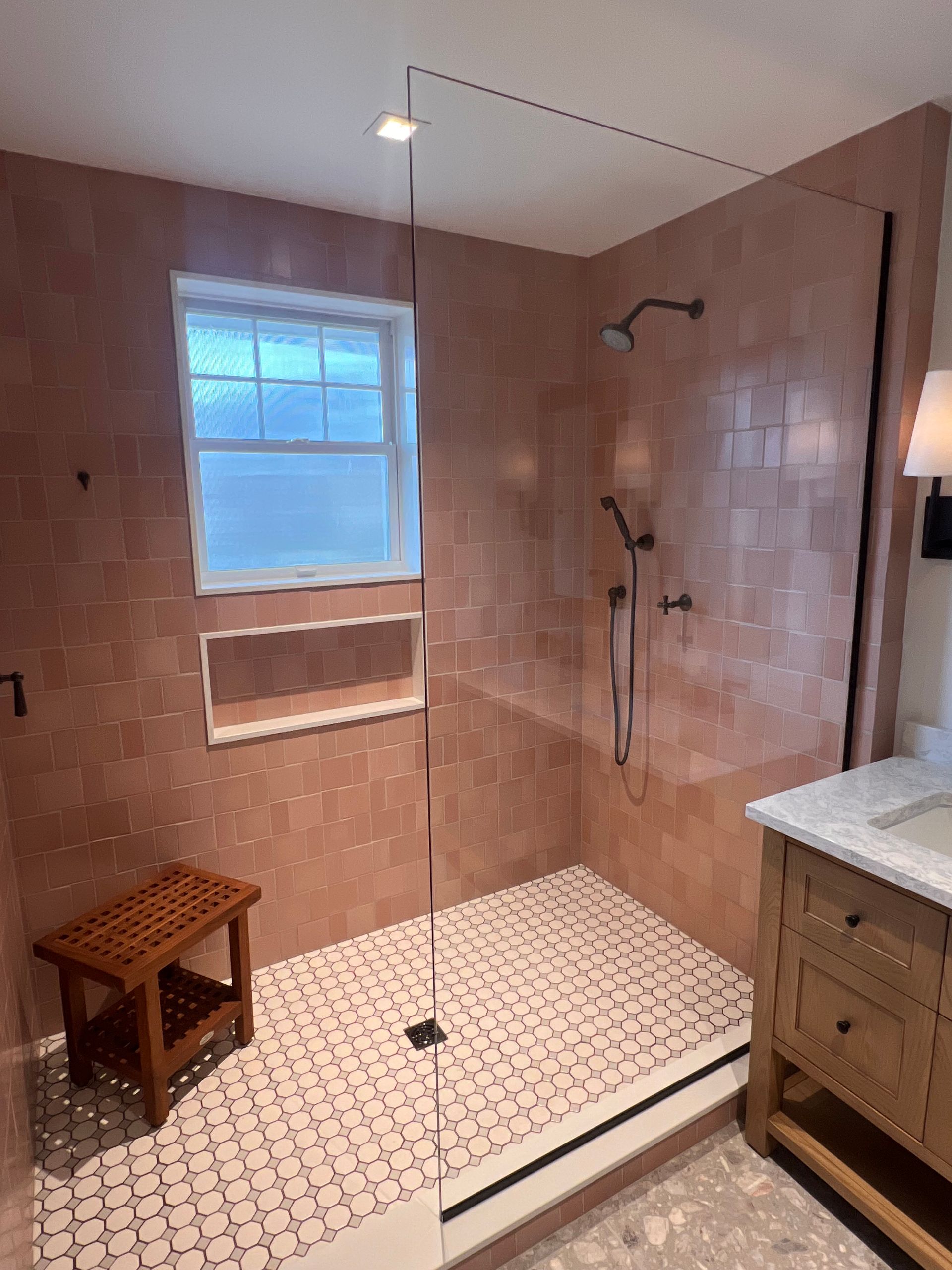 A walk-in shower with rose-colored wall tiles, patterned tile floors, a glass panel, and a small wooden stool inside.