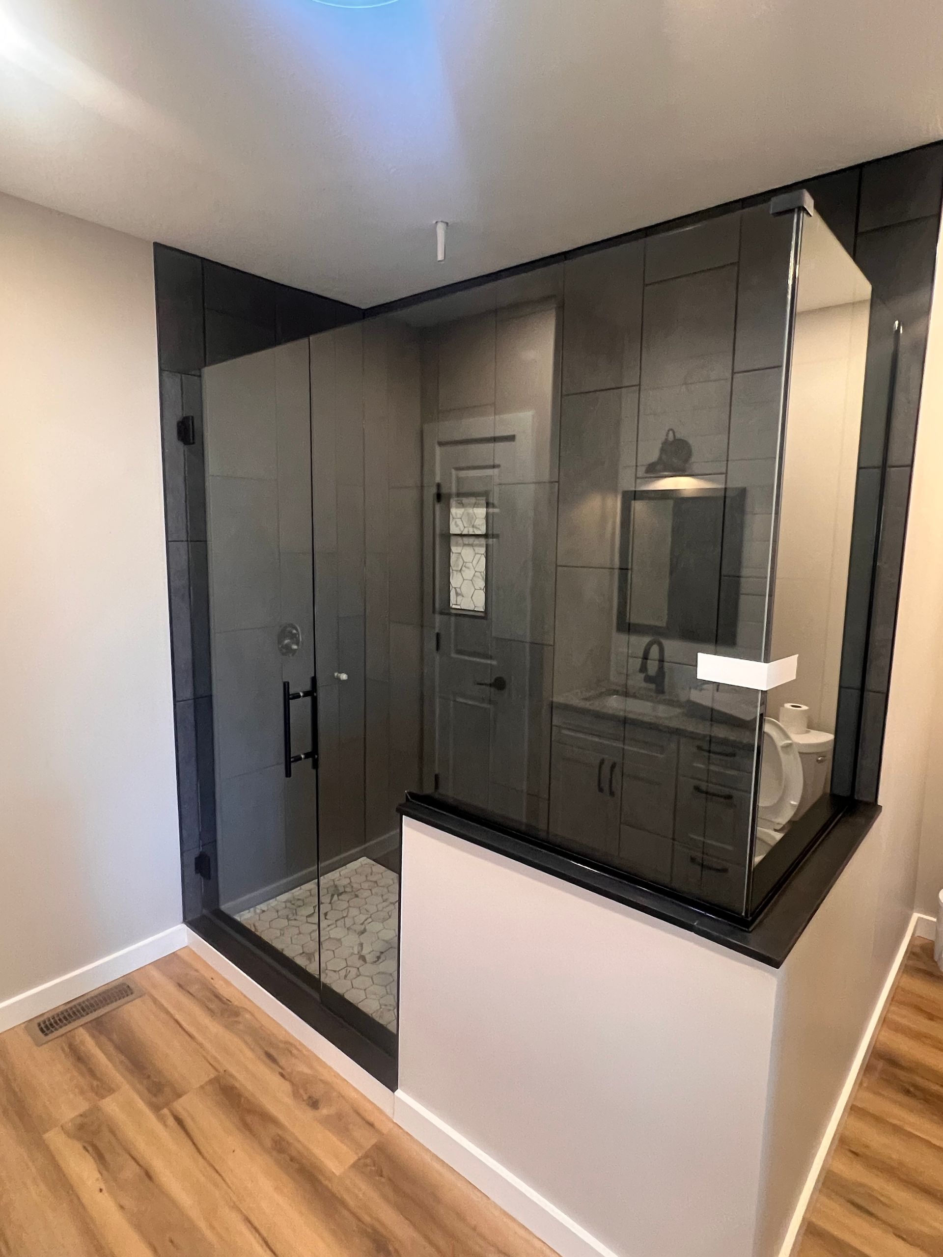 A modern shower with glass walls, black trim, and dark tile, situated next to a white vanity in a bathroom with wood floors.