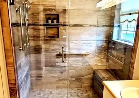 A walk-in shower with brown stone-look tile walls, a built-in bench, glass door, grab bars, and a window.