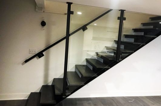 Black staircase with glass railing, white walls, and gray floor.