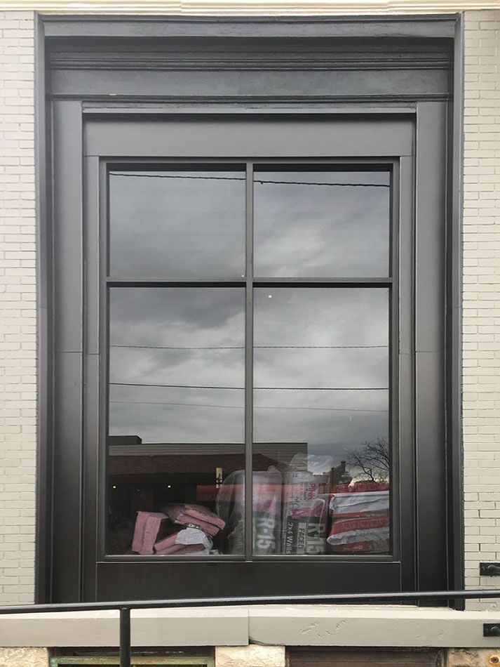 Large, dark framed window set in light brick wall. Reflections of sky, buildings and store goods inside.
