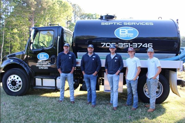 Five men standing beside a black Septic Services tanker truck in a grassy outdoor area.