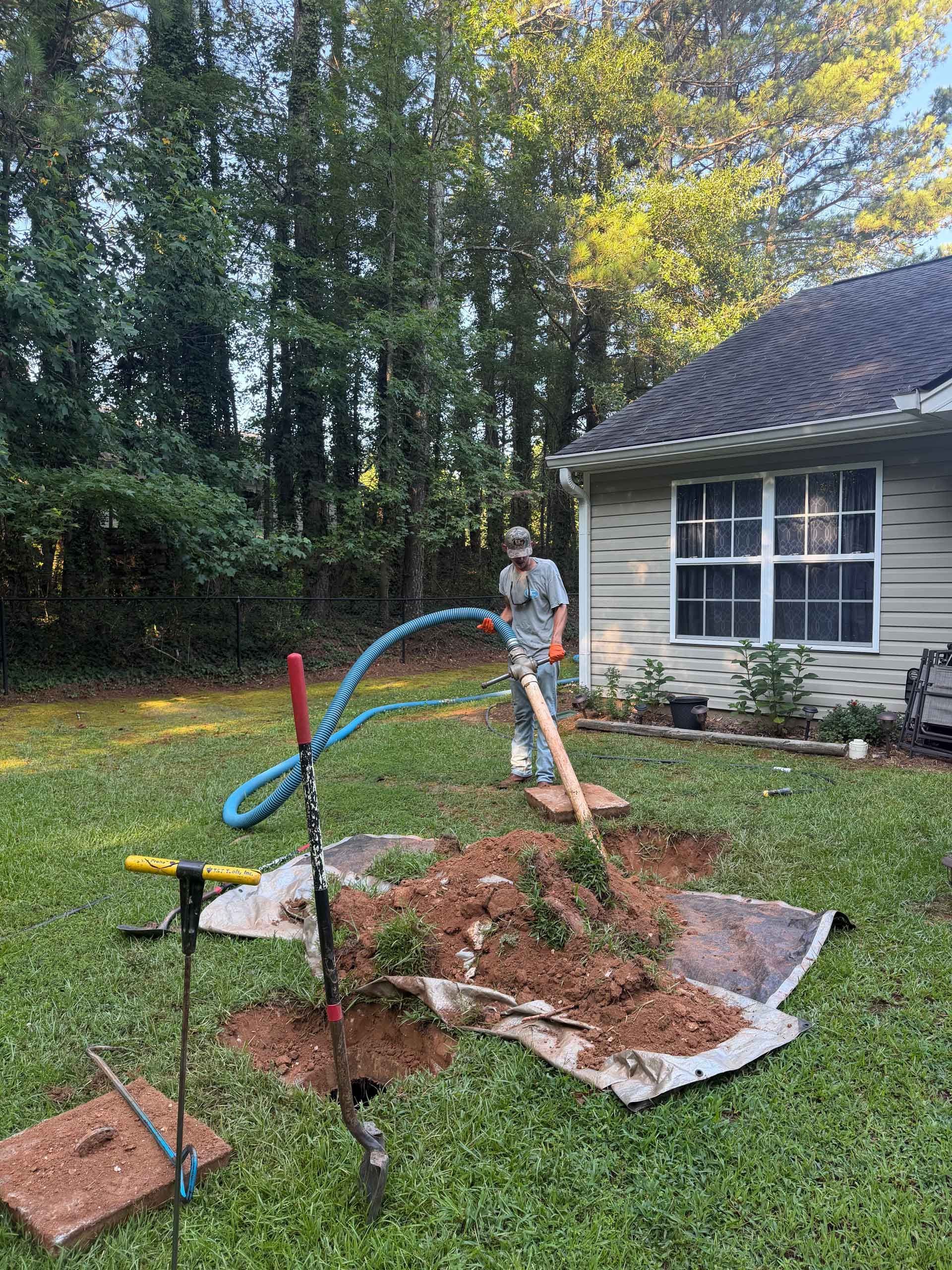 Person watering a garden bed beside a house with a blue hose and freshly turned soil
