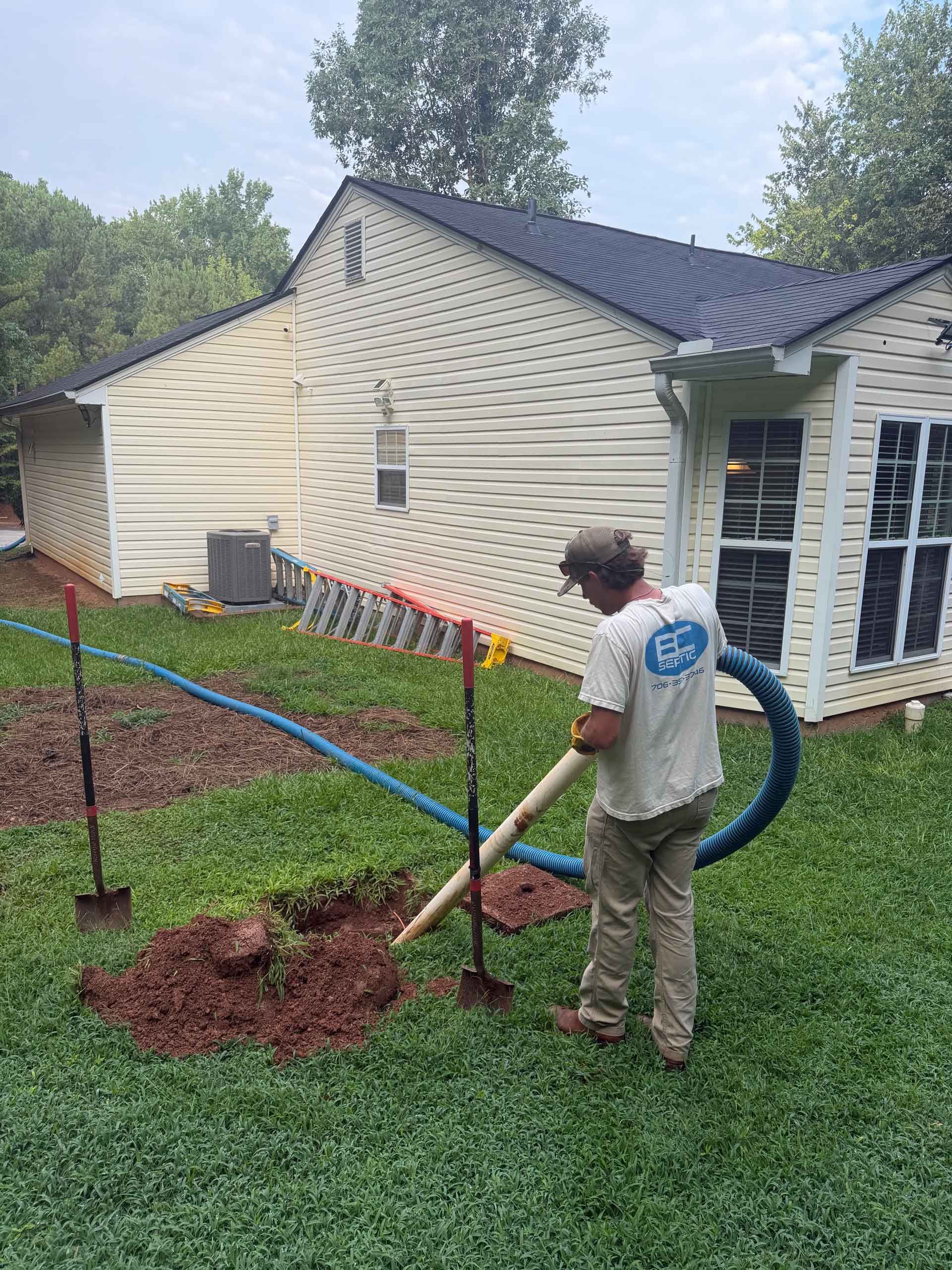 Worker uses a hose vacuum to mulch garden beds beside a beige house