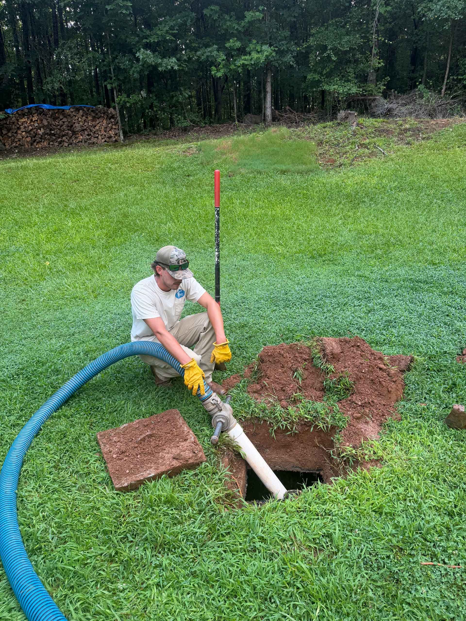 Worker cleaning a drainage hole in a grassy yard, using a hose and tools near a covered utility box