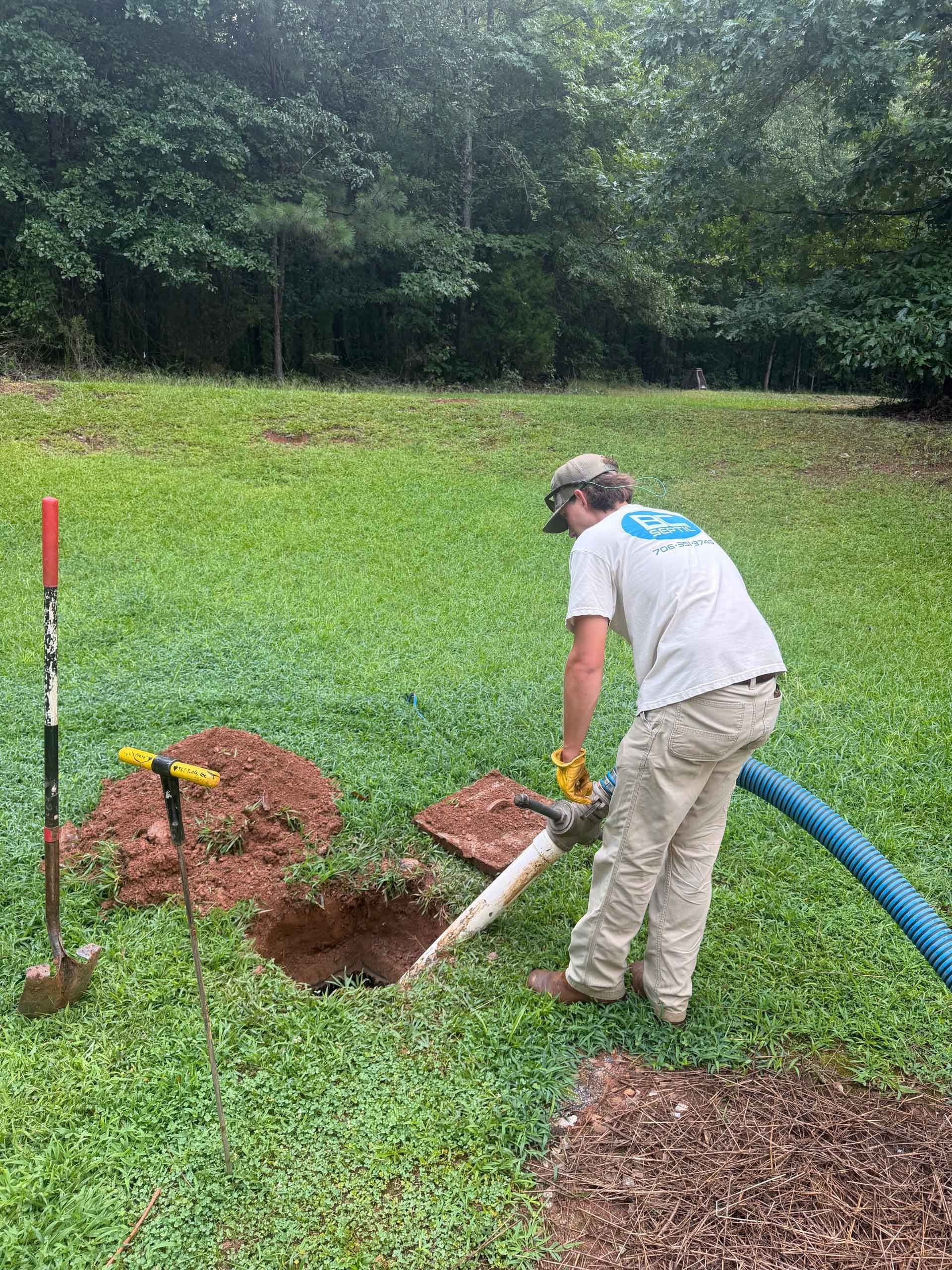 Person in beige clothes uses a white and blue hose to spray soil in a grassy yard near a small dirt pit.