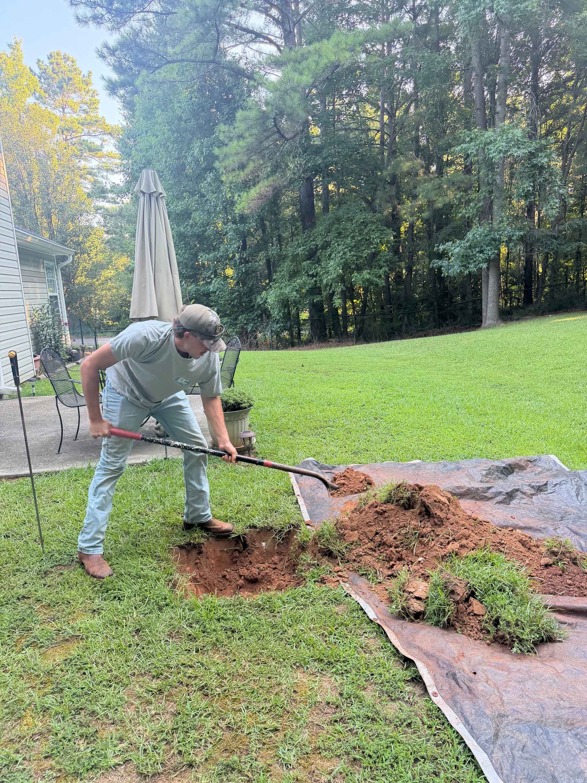 Person raking mulch beside a lawn in a backyard with trees and a patio umbrella