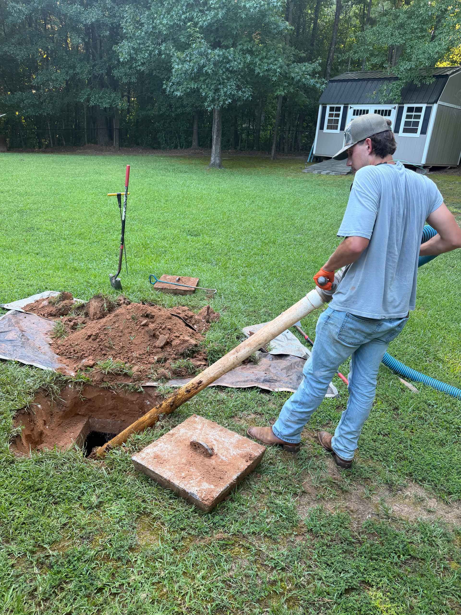 Worker using a long tool beside a dug hole in a grassy yard with a hose and shed nearby