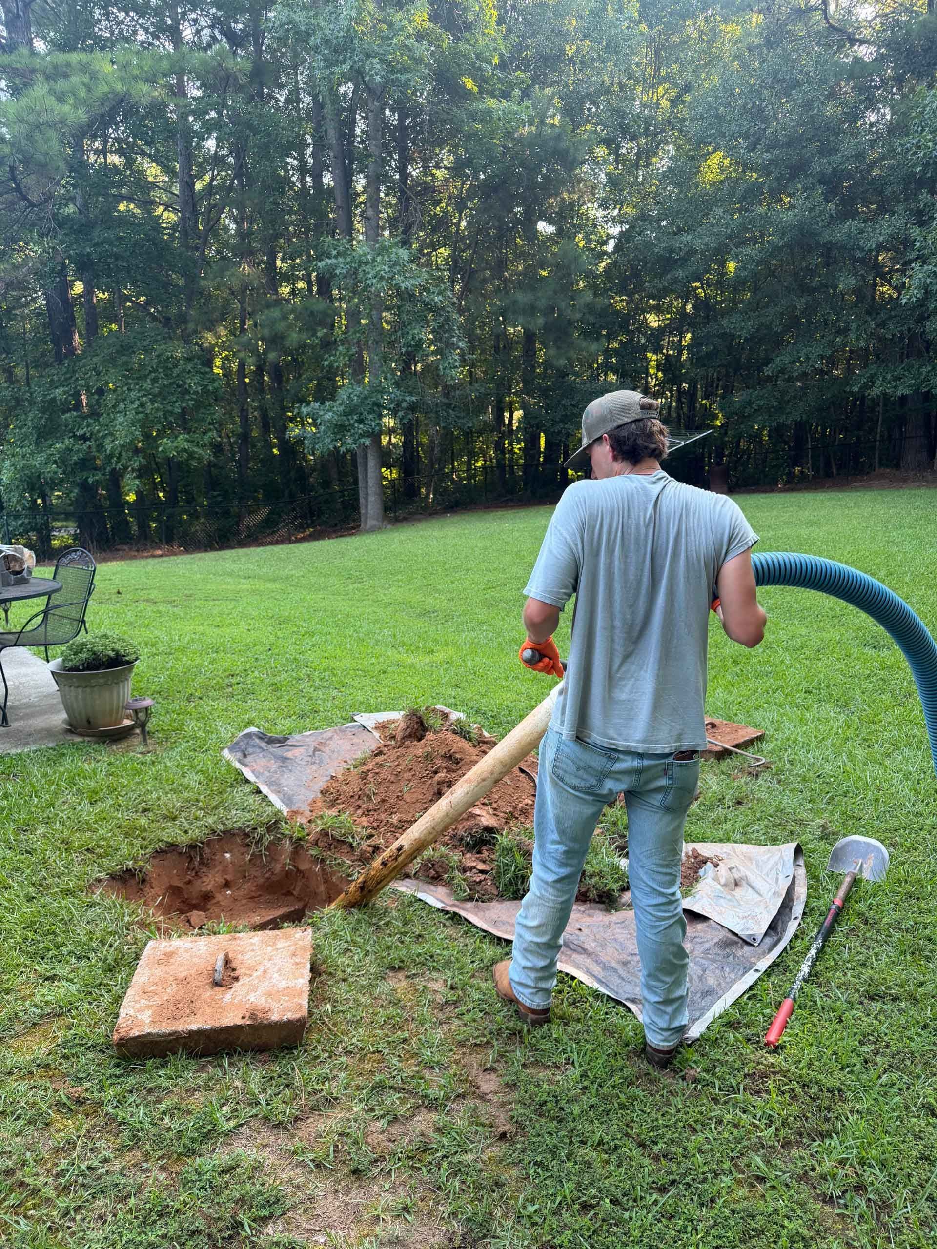 Person digging a hole in a grassy backyard beside a tarp and tools, with trees in the background