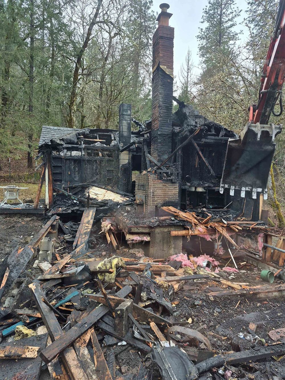 Charred remains of a house after a fire, with debris and excavator arm visible.