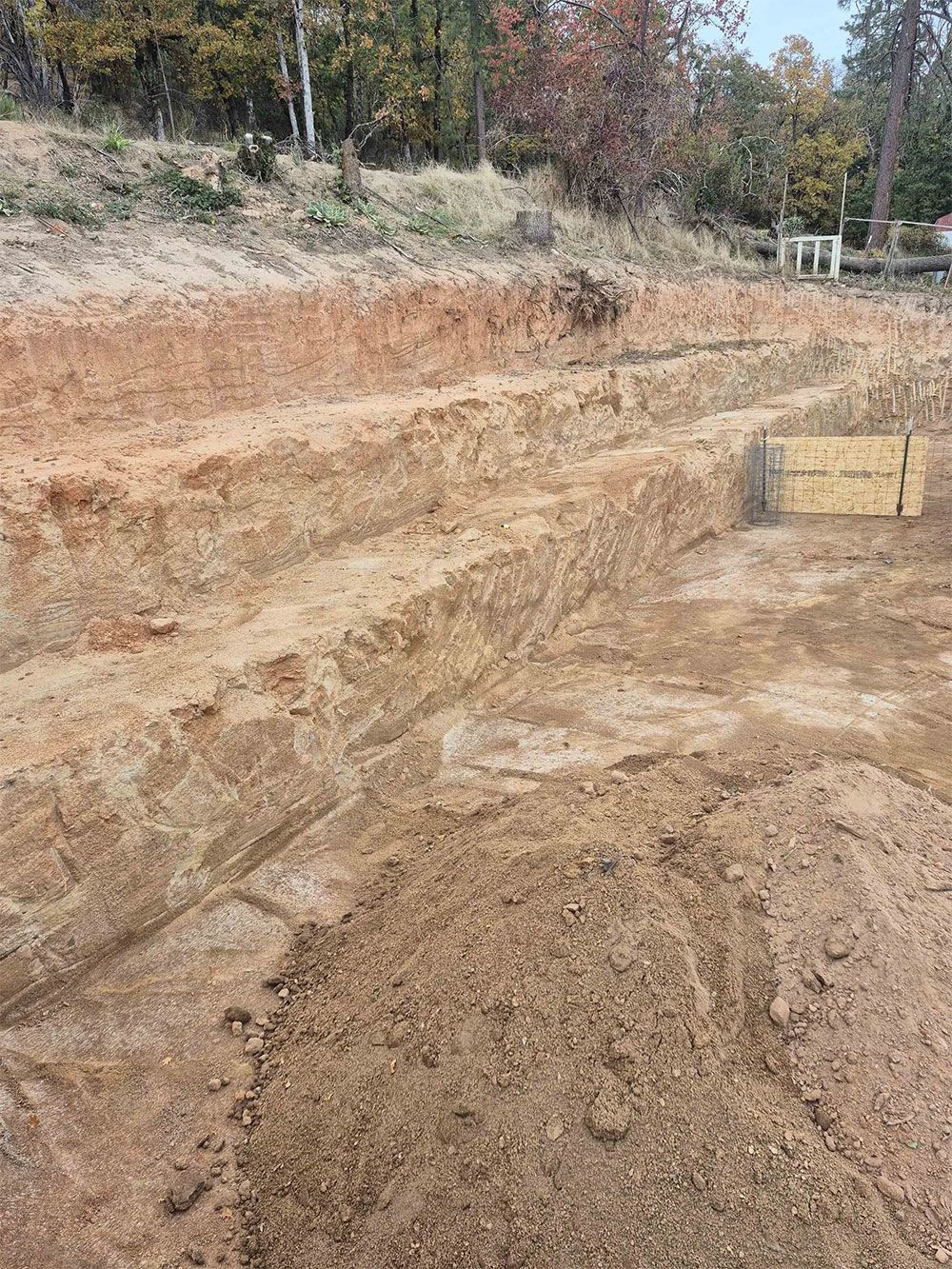 Excavation site with rectangular trenches dug into reddish-brown earth.  Forest and blue sky visible in the background.