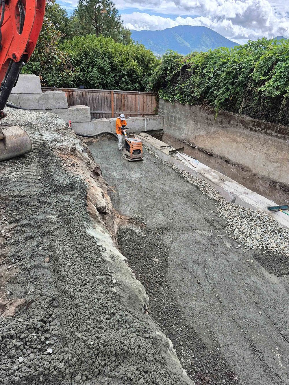 Construction worker compacts gravel with a machine, near a concrete wall. Mountain visible in background.