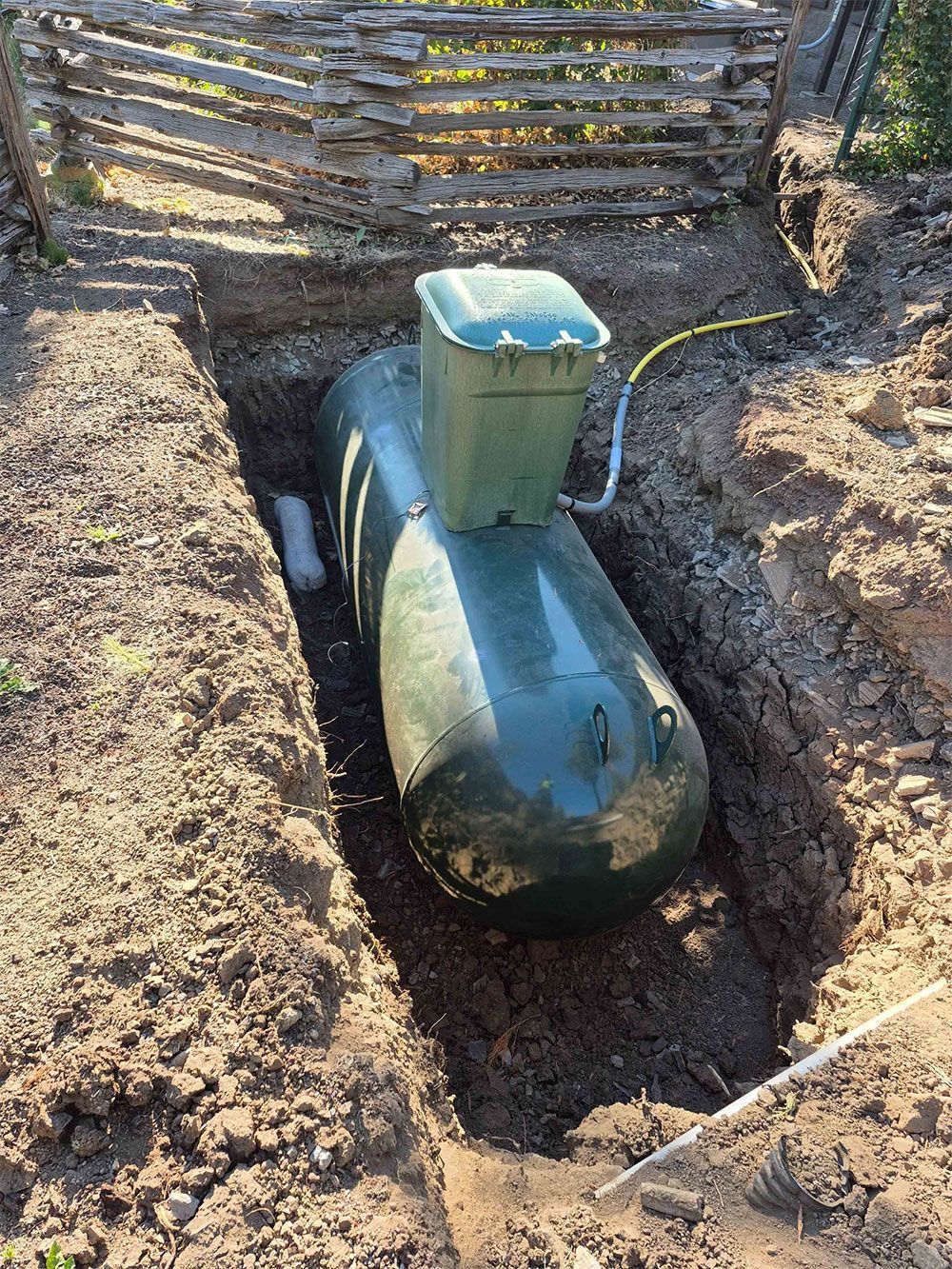 An elongated green tank installed in a dirt trench, near a wooden fence.